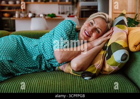 Reife Frau in einem blaugrünen Kleid, die auf einer grünen Couch liegt und ein buntes Kissen umschließt. Das gemütliche Wohnzimmer bietet Entspannung und Komfort in Stockfoto