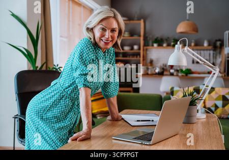 Glückliche, reife Geschäftsfrau, die einen Laptop benutzt und von zu Hause aus arbeitet, an einem Holztisch in der gemütlichen Küche sitzt und freiberuflich online arbeitet Stockfoto