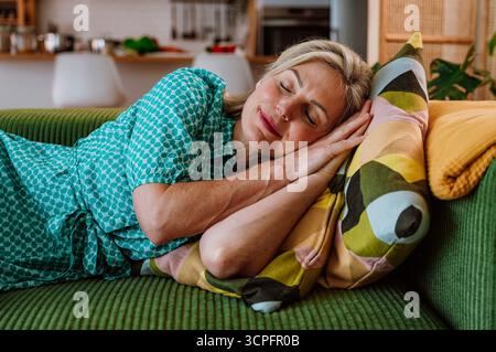 Reife Frau in einem blaugrünen Kleid, die auf einer grünen Couch liegt und ein buntes Kissen umschließt. Das gemütliche Wohnzimmer bietet Entspannung und Komfort in Stockfoto