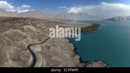 Der Blick auf eine gewundene Straße schlängelt sich durch die trockene Landschaft entlang des türkisfarbenen Baisha Lake, im Kontrast zu den schneebedeckten Bergen in der Ferne, Baisha Lake, Xinjiang, China. Stockfoto