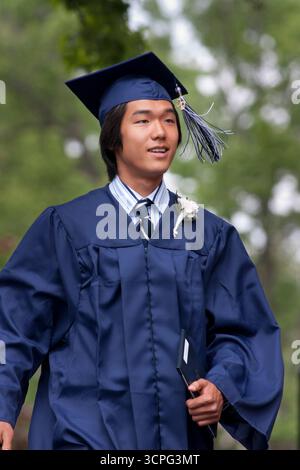 Ein junger Schüler geht selbstbewusst in Mütze und Kleid, nachdem er sein Diplom bei seiner Abschlussfeier erhalten hat. Stockfoto