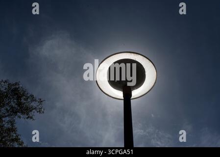 Scheibenförmige Laternenpfosten zwischen Kiefern, ausgeschaltet und mit der Sonne dahinter, die den Effekt erzeugt, an einem bewölkten Tag eingeschaltet zu werden. Stockfoto