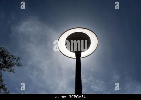Scheibenförmige Laternenpfosten zwischen Kiefern, ausgeschaltet und mit der Sonne dahinter, die den Effekt erzeugt, an einem bewölkten Tag eingeschaltet zu werden. Stockfoto