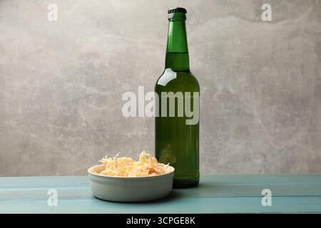 Getrocknete Tintenfischspäne und Flasche Bier auf hellblauem Holztisch vor grauem Hintergrund, Nahaufnahme Stockfoto