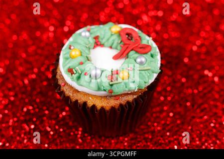 Festlicher Cupcake mit grünem Glaskränz und bunten Streuseln auf einem leuchtend roten Glitzerhintergrund Stockfoto
