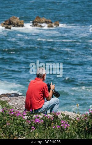 Ein Tourist mit grauen Haaren sitzt am Meer mit einer Kamera in der Hand. Stockfoto