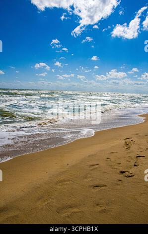 Eine kraftvolle und stimmungsvolle Strandszene. Schaumige Wellen krachen auf einen breiten, orangen Sandstrand unter einem dramatischen, tiefblauen Himmel voller verstreuter weißer C Stockfoto