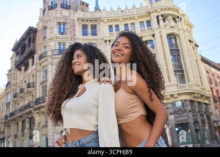 Zwei fröhliche junge Frauen mit lockigen Haaren lächeln und stehen während eines Städtetrips vor der historischen europäischen Architektur Stockfoto