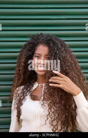 Fröhliches Porträt der jungen Frau, das fröhlich lächelt und natürliches langes, lockiges braunes Haar vor einem grünen Hintergrund zeigt Stockfoto