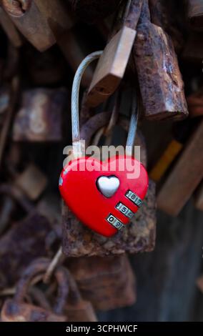 Nahaufnahme eines romantischen Vorhängeschlosses aus dem Herzen eines roten Liebhabers, das mit anderen alten, rostigen Vorhängeschlössern auf einer Brücke verriegelt ist Stockfoto