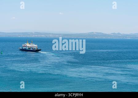 Eine Fähre gleitet über das ruhige blaue Wasser und hinterlässt ein wahres Licht, während sie an einem sonnigen Tag zu weit entfernten Hügeln im Hintergrund fährt. Der Himmel ist brig Stockfoto