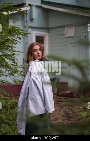 Ein Nahporträt einer Frau in einem grünen Kleid mit einer Jeansjacke über den Schultern, die neben einem pastellfarbenen Holzhaus steht Stockfoto