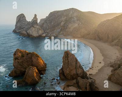 Blick aus der Vogelperspektive auf dramatische Klippen und goldenen Sand treffen auf das türkisfarbene Meer am Praia da Ursa, dem westlichsten Strand Europas, eine Symphonie aus Licht und Schatten Stockfoto