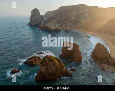 Aus der Vogelperspektive treffen auf zerklüftete Klippen auf das türkisfarbene Meer, Wellen, die unter dem goldenen Licht gegen die Felsen krachen, Praia da Ursa, Lissabon, Portugal. Stockfoto