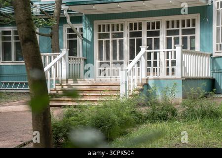 Ein Blick aus der Nähe auf den hölzernen Eingang eines historischen Hauses in Peredelkino zeigt weiße Geländer, breite Stufen und türkisfarbene Wände, die von Grün eingerahmt sind Stockfoto