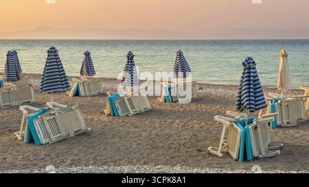 Leere Liegestühle und zusammengefaltete Sonnenschirme säumen einen Sandstrand, an einem ruhigen Meer Stockfoto