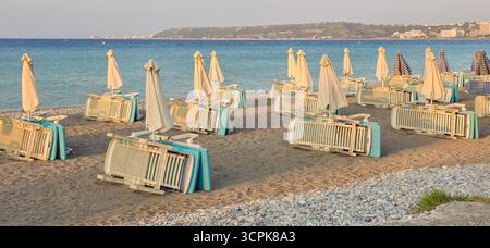 Leere Liegestühle und zusammengefaltete Sonnenschirme säumen einen Sandstrand, an einem ruhigen Meer Stockfoto