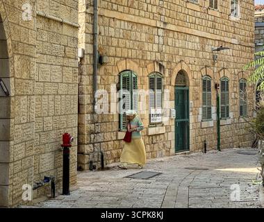 Jerusalem, Israel - 30. Oktober 2019: Blick auf eine Frau in traditioneller Kleidung, die entlang einer gepflasterten Straße geht, die von Gebäuden aus Jerusalem-Stein eingerahmt ist, deren Texturen durch Sonnenlicht erwärmt werden. Stockfoto