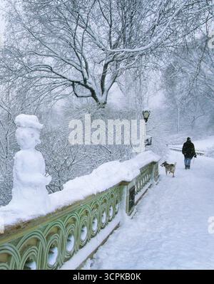 New York Bow Bridge Schnee. Schneemann und Mann Wanderhund. Central Park in Manhattan, New York City, USA. Bäume mit frischem Schnee. Baujahr 1860 Stockfoto