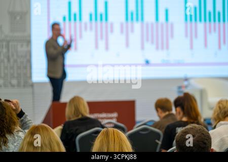 Der Moderator auf der Bühne erklärt Marktdaten, während das Publikum einen großen Bildschirm mit bunten Balken und Linien ansieht. Professionelles Umfeld, das sich auf geschäftliche Erkenntnisse und Trends konzentriert Stockfoto