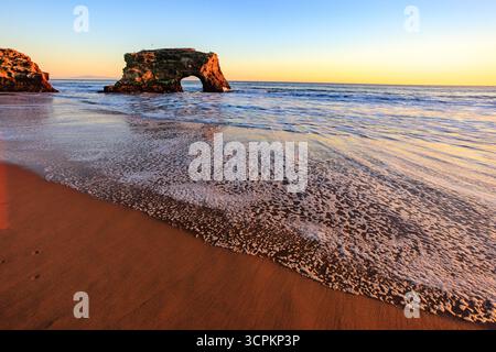 Ein malerischer Blick auf den Natural Bridges State Beach in Santa Cruz, Kalifornien, mit der berühmten natürlichen Brückenformation während eines pulsierenden Sonnenuntergangs. Das Bild fängt die ruhige Schönheit des Strandes mit sanften Wellen an Land ein, die das Naturwunder und die ruhige Atmosphäre der Küstenlandschaft hervorheben. Stockfoto