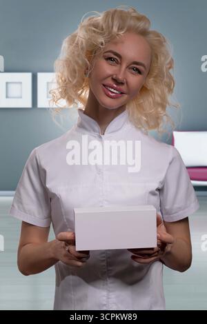 Blonde Frau in einem weißen medizinischen Mantel lächelt, während sie ein Zahnaufhellungspaket in einer hellen modernen Klinik hält. Sie wirkt freundlich und professionell. Stockfoto