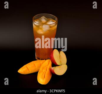 Apfel-Mangosaft in einem hohen facettierten Glas mit Eis auf schwarzem Hintergrund, neben reifem Obst. Nahaufnahme. Stockfoto