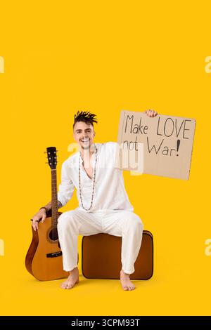 Männlicher Hippie, der Karton mit Text hält, MACHT LIEBE NICHT KRIEG und Gitarre auf gelbem Hintergrund Stockfoto