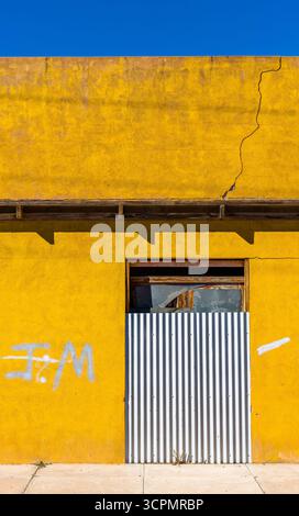 Eine leuchtend gelbe Wand eines verlassenen Gebäudes wird vor einem klaren blauen Himmel gezeigt. Die Wand weist sichtbare Risse und Graffiti in weiß auf. Stockfoto