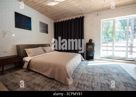 Modernes Schlafzimmer mit minimalistischem Design, großem Bett, weißen Ziegelwänden, dunklen Vorhängen und einem Fenster mit Blick auf die Natur. Stockfoto