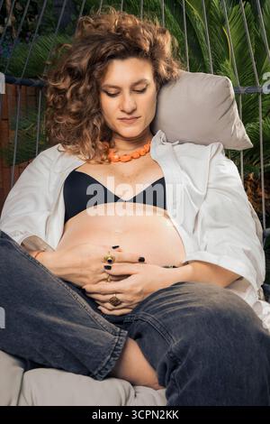 Eine nachdenkliche schwangere Frau mit lockigen Haaren sitzt allein in einem hängenden Stuhl im Freien und hält ihr Baby sanft in einem ruhigen Moment der Introspektion und Stockfoto