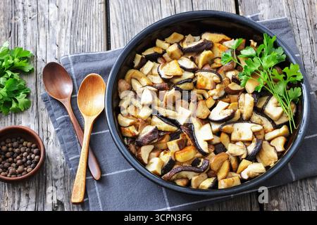 Gebratene Wildpilze, Ceps in einer schwarzen Schüssel auf einem rustikalen Holztisch mit Holzlöffeln, Allspice Beeren und Petersilie, horizontaler Blick von oben Stockfoto