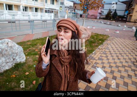Elegante kaukasische Dame in klassischer Oberbekleidung schlendert durch die Stadt und führt ein Telefongespräch. Eleganter Stadtspaziergang und digitale Kommunikation Stockfoto