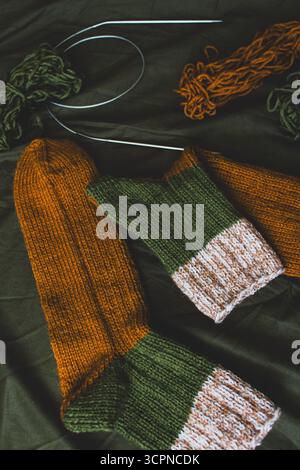Kuschelige Stricksocken mit Garnknäuel und Stricknadeln. Gestreifte Wollsocken auf dunkelgrünem Laken. Kuschelige Herbstfarben. Gewebtes Häkeln aus nächster Nähe. Stockfoto