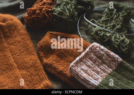 Kuschelige Stricksocken mit Garnknäuel und Stricknadeln. Gestreifte Wollsocken auf dunkelgrünem Laken. Kuschelige Herbstfarben. Gewebtes Häkeln aus nächster Nähe. Stockfoto