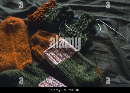 Kuschelige Stricksocken mit Garnknäuel und Stricknadeln. Gestreifte Wollsocken auf dunkelgrünem Laken. Kuschelige Herbstfarben. Gewebtes Häkeln aus nächster Nähe. Stockfoto