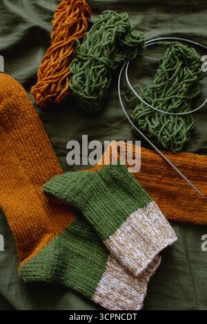 Kuschelige Stricksocken mit Garnknäuel und Stricknadeln. Gestreifte Wollsocken auf dunkelgrünem Laken. Kuschelige Herbstfarben. Gewebtes Häkeln aus nächster Nähe. Stockfoto