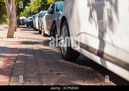 Mehrere Autos parken auf einem gepflegten, gepflasterten Bürgersteig. Sonnenlicht filtert durch Bäume und wirft Schatten auf den Boden. Diese Einstellung erfasst einen Stockfoto
