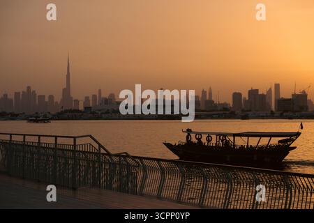 Dubai Creek Harbour Uferpromenade mit atemberaubendem Blick auf die Skyline der Stadt und den berühmten Burj Khalifa mit Boot im Bach, Silhouette bei Sonnenuntergang Stockfoto