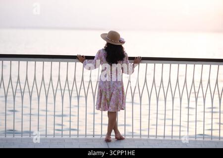 Junge Frau, die auf der Uferpromenade mit Blick auf die Skyline von Dubai bei Sonnenuntergang steht, Blick vom Hafen des Dubai Creek, sie trägt ein Blumenkleid Stockfoto