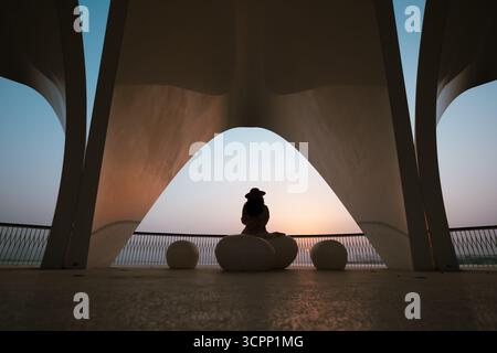 Touristenfrau mit Hut und fließendem Kleid, die den Sonnenuntergang mit Blick auf die Skyline in Dubai beobachtet, Blick von der Hafenpromenade des Dubai Creek Stockfoto