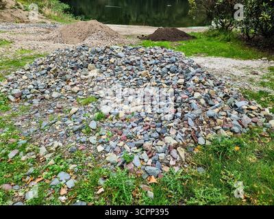 Großer Haufen mehrfarbiger Kies und kleinere Erdhaufen auf dem Boden in der Nähe von Gras, die sich auf die Landschaftsgestaltung am Wasser vorbereiten Stockfoto