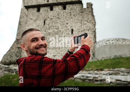 Fröhlicher junger Mann lächelt, während er eine alte Steinfestung Vrsac in Serbien fotografiert. Reise-Blogger, der Videos im Freien macht. Stockfoto