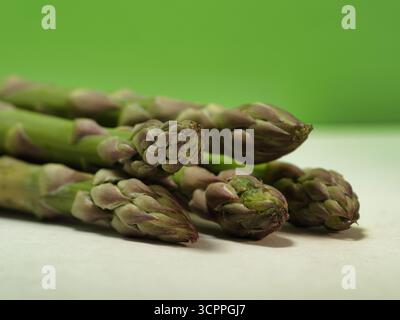 Fresh Green Spargelspitzen Nahaufnahme – Makroaufnahme von rohen grünen Spargelspitzen vor einem leuchtend kalkgrünen Hintergrund und weißer Oberfläche. Konzept für Stockfoto