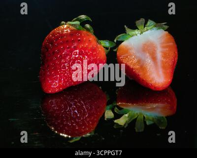 Erdbeerhalbierung auf schwarzem Spiegel - zwei reife, rote Erdbeeren, eine ganze und eine in zwei Hälften geschnitten, reflektiert auf einer glänzend schwarzen Oberfläche. Moody Stockfoto