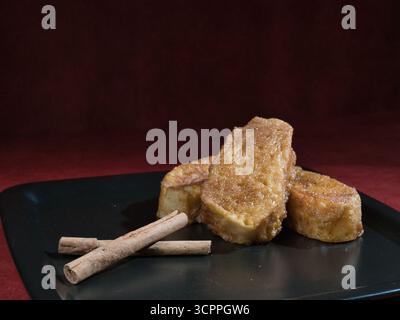 Zimt French Toast Dessert Dark – drei Scheiben süßen French Toast (Torrijas) mit Zimt und Zucker auf einem schwarzen Teller mit Zimtstangen. Dunkel Stockfoto