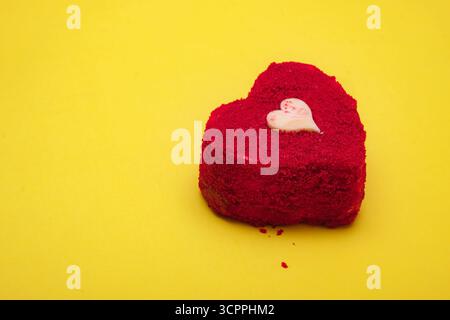 Herzförmiger roter Samtkuchen mit einem kleinen weißen Schokoladenherz auf gelbem Hintergrund. Liebe, Romantik, Valentinstag. Stockfoto