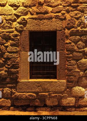 Eine rustikale Steinmauer mit einem alten Gitterfenster wird nachts durch warmes goldenes Licht beleuchtet. Ideal für Themen im Zusammenhang mit der Geschichte. Stockfoto