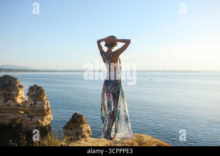 Nicht wiedererkennbare Frau in einem langen Sommerblumenkleid, die mit erhobenen Armen an die Kamera zurücksteht und die Haare berührt und wunderschöne Felsformationen beobachtet. Stockfoto