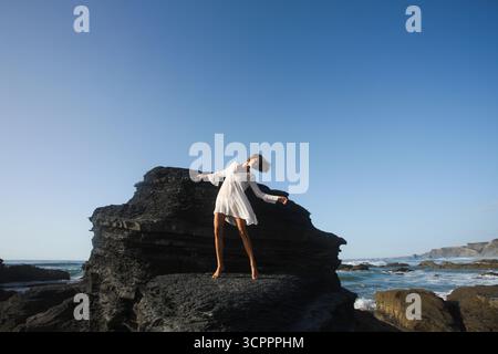 Schöne natürliche Frau in romantischem schlichtem weißem Kleid, die ihre Schönheit genießt, während sie eine elegante Back Band tanzt vor dunklen, einzigartigen Felsen. Stockfoto